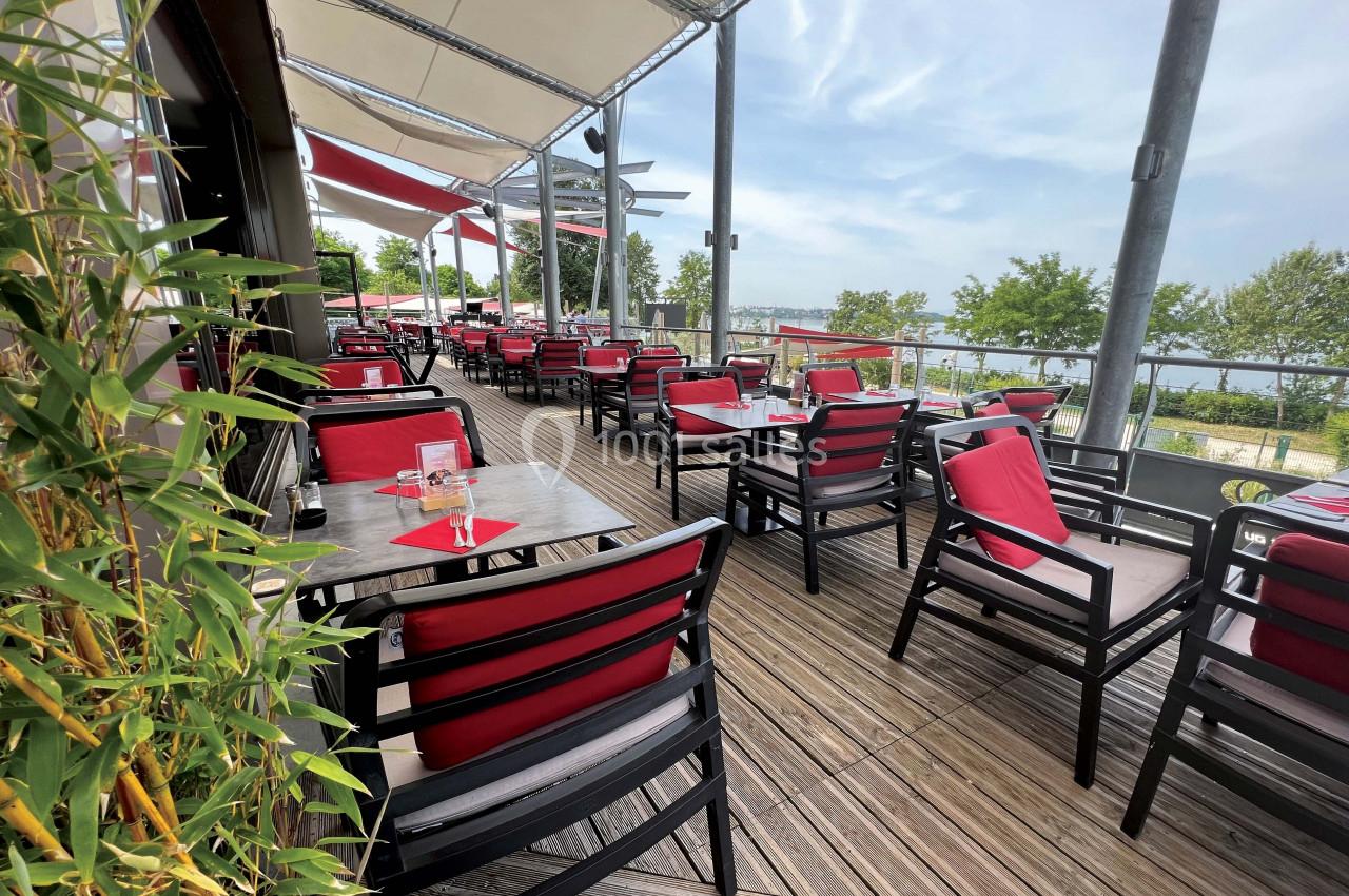 Terrasse d'un restaurant avec des tables et chaises rouges, vue sur un paysage arboré et un plan d'eau.