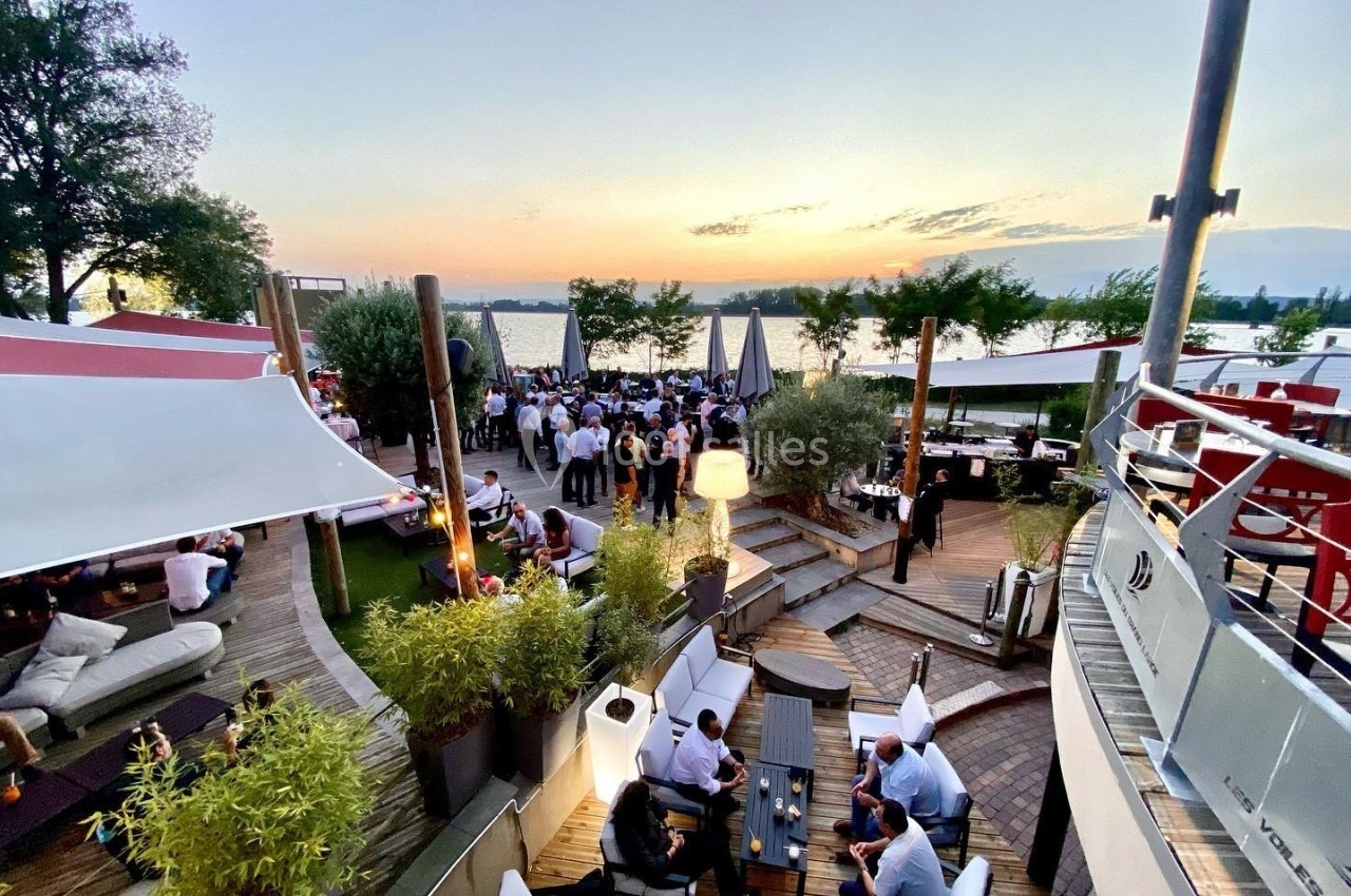Terrasse animée au bord d'un lac au coucher du soleil, avec des groupes de personnes assises et debout.