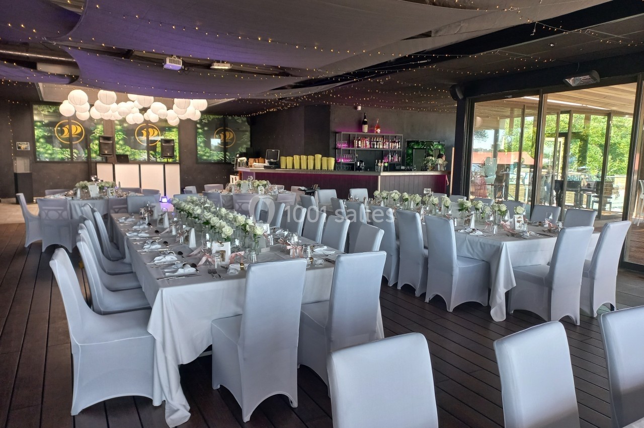 Salle de réception décorée avec des tables alignées, nappes blanches, chaises houssées et éclairage tamisé.
