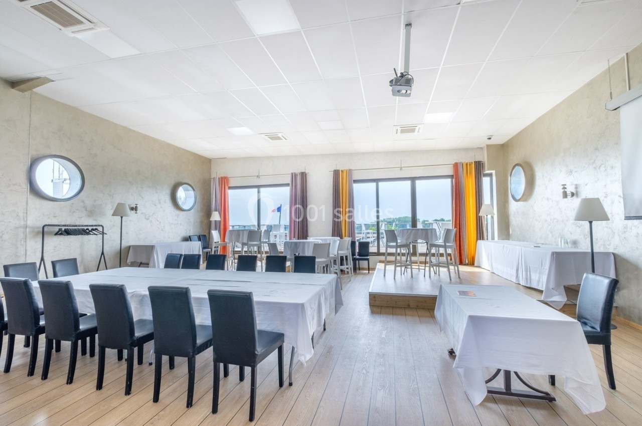 Salle lumineuse avec grandes fenêtres, tables recouvertes de nappes blanches et chaises noires, décor sobre et épuré.