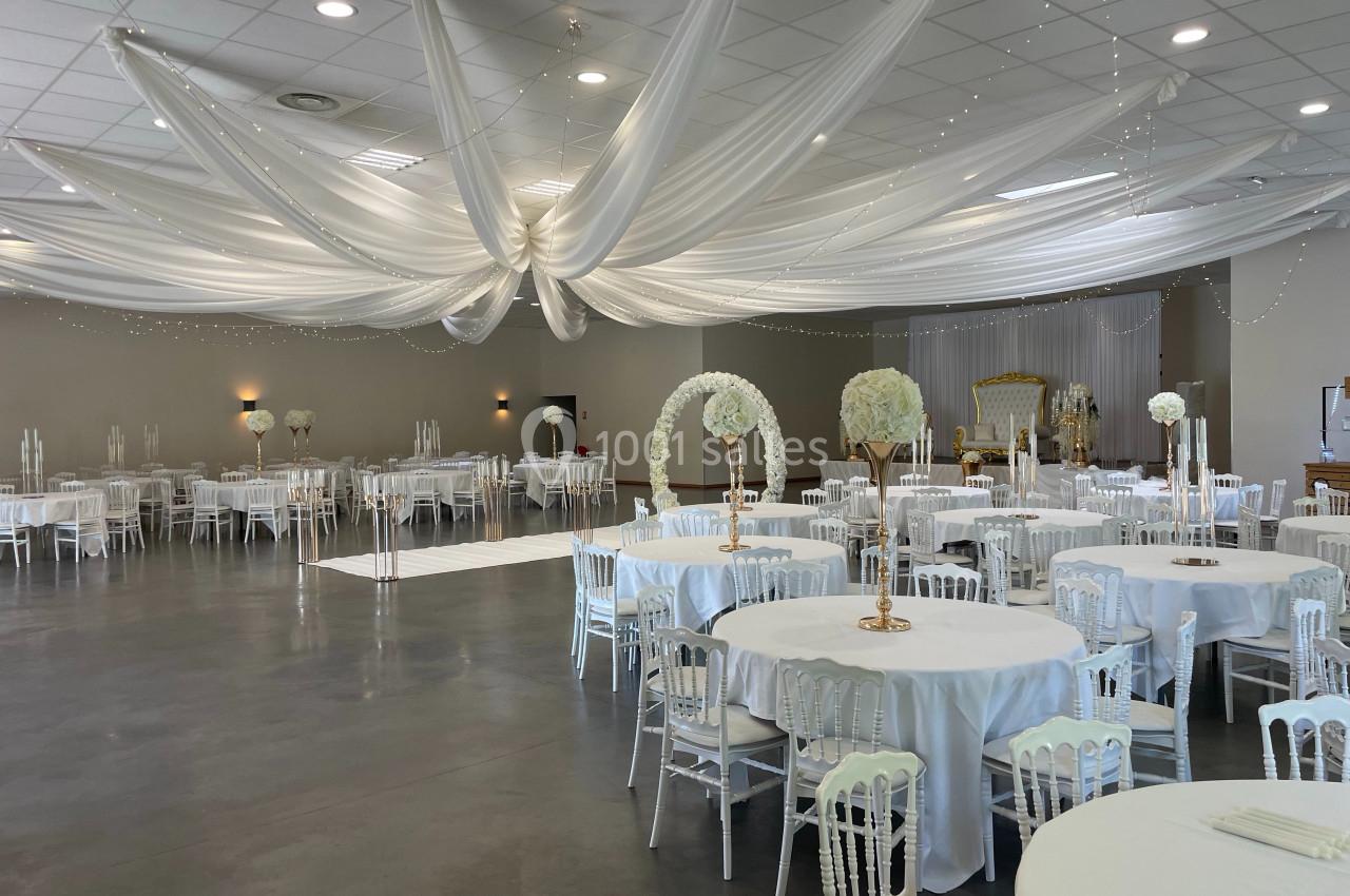 Salle de réception décorée avec des tables rondes, des nappes blanches, des chaises assorties et un plafond drapé de tissu…