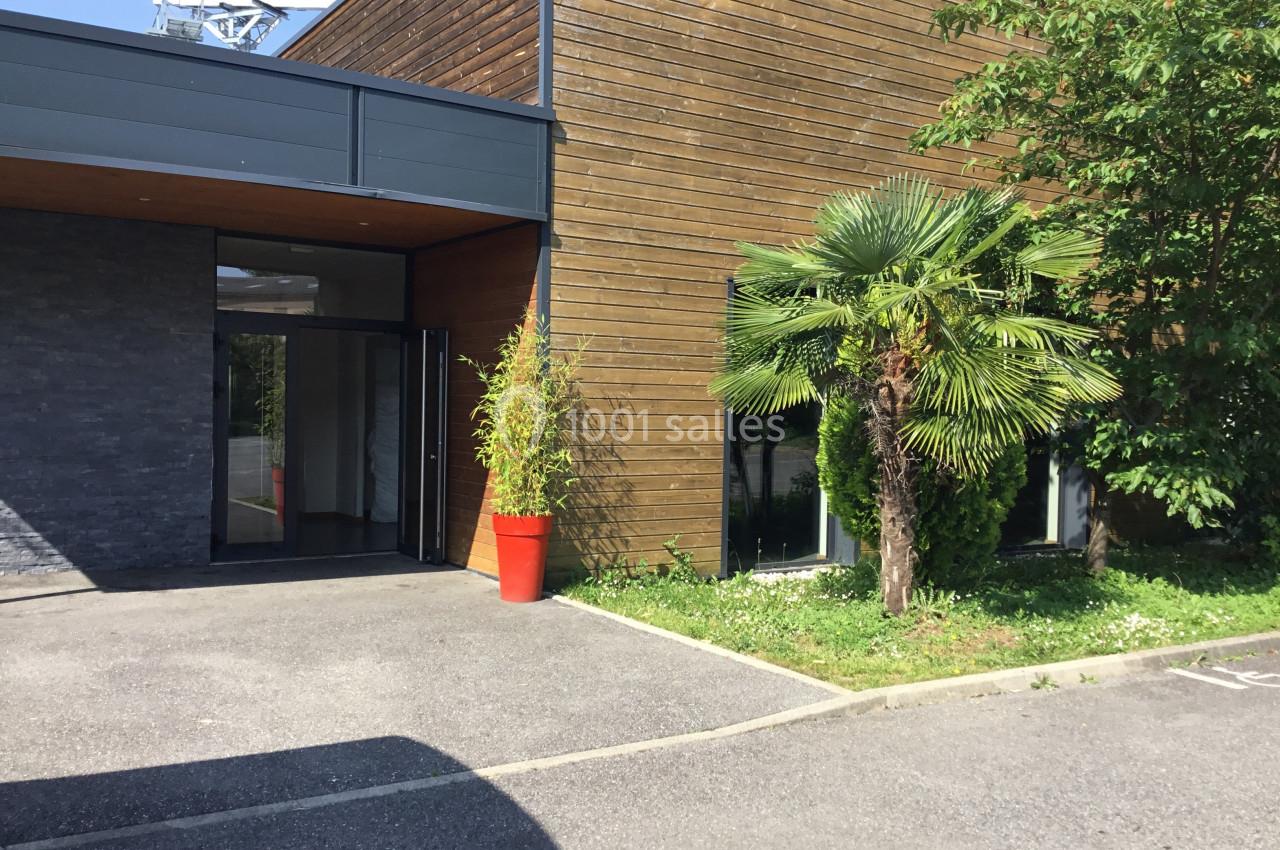 Entrée d'un bâtiment moderne avec façade en bois, plantes vertes et un pot rouge près de la porte vitrée.