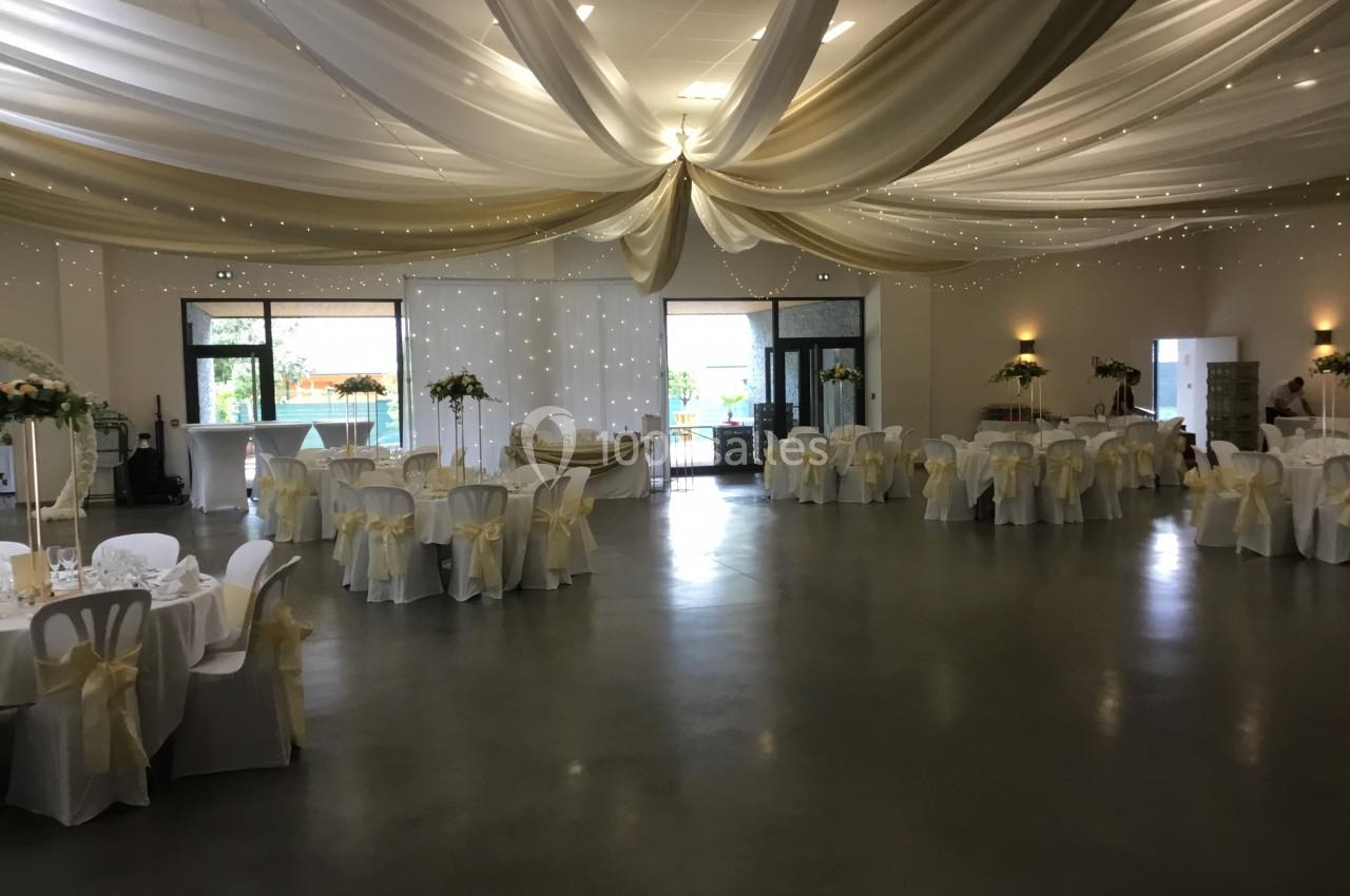 Salle de réception décorée avec des tables rondes, nappes blanches, chaises ornées de nœuds et drapés au plafond.