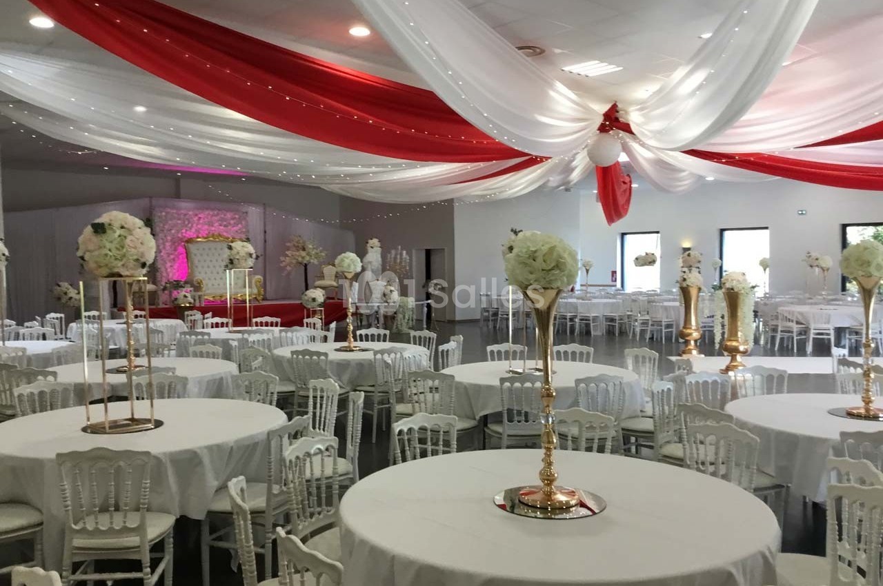 Salle de réception décorée avec des tables rondes, des chaises blanches et des drapés rouges et blancs au plafond.
