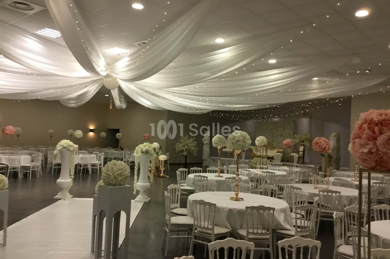 Salle de réception décorée avec des tables rondes, des chaises blanches et des drapés blancs suspendus au plafond.
