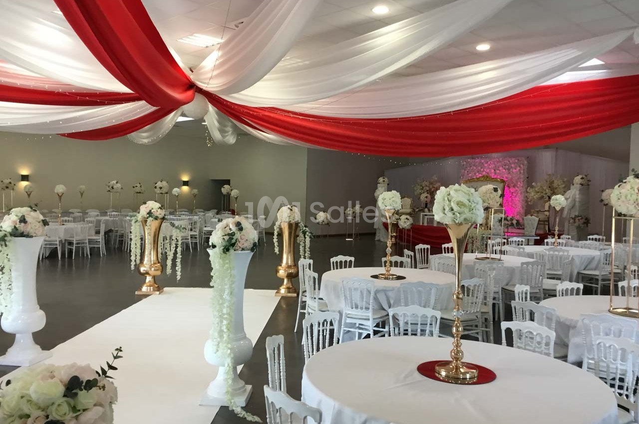 Salle de réception décorée avec des drapés rouges et blancs au plafond, tables rondes et arrangements floraux élégants.