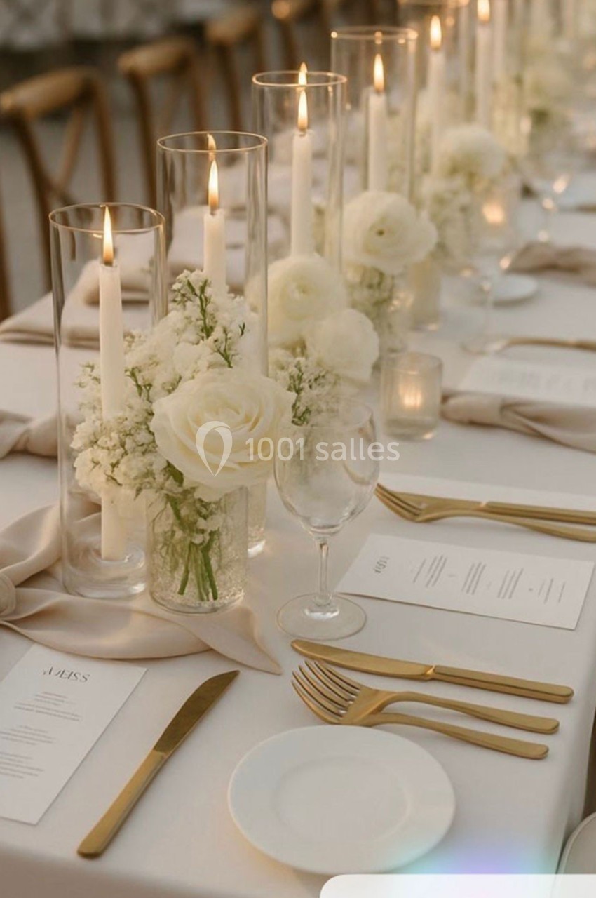 Table élégamment dressée avec bougies allumées, fleurs blanches et couverts dorés sur une nappe blanche.
