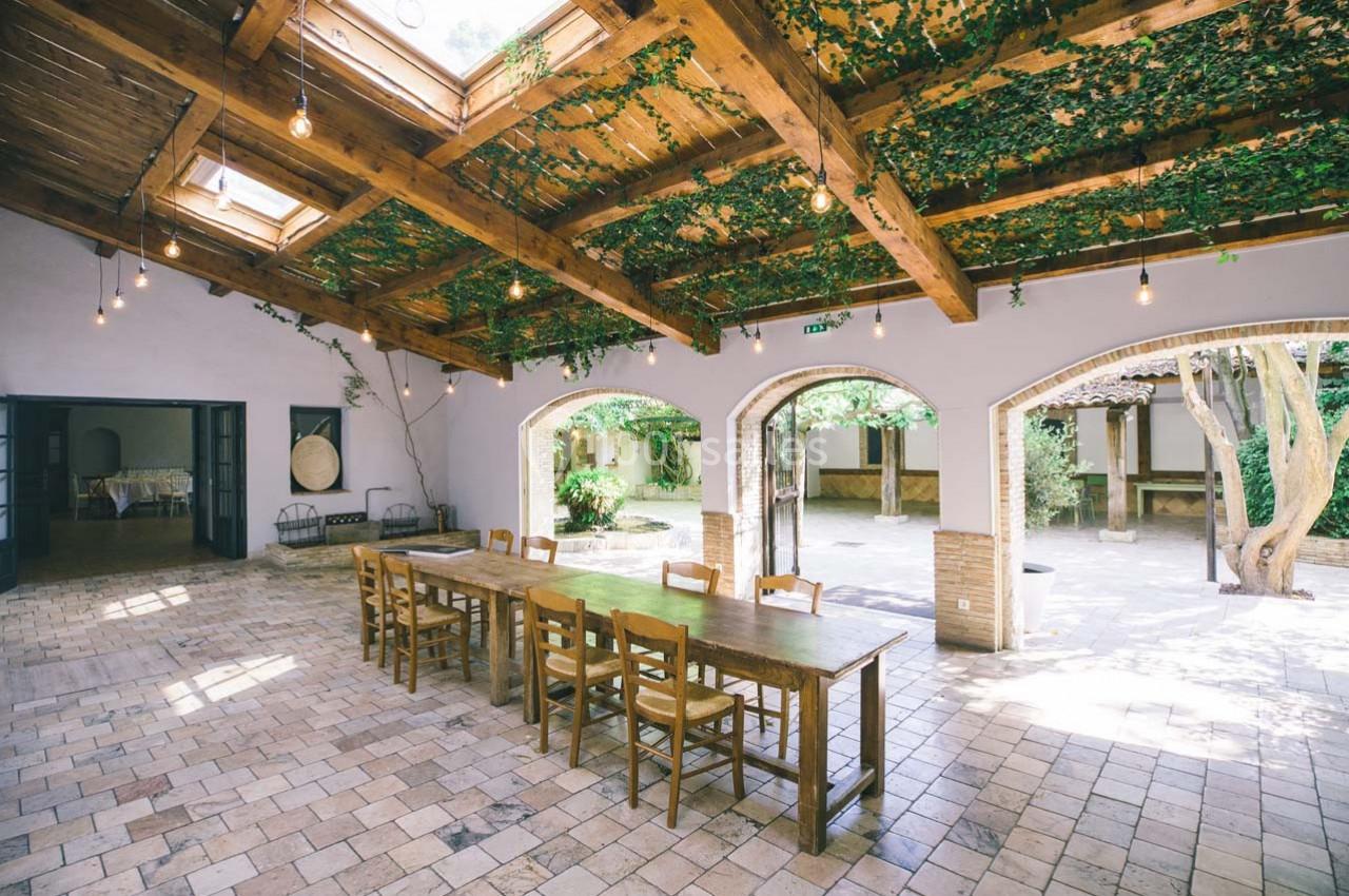 Location salle Gémenos (Bouches-du-Rhône) - Moulin de Gemenos #2 Salle lumineuse avec toit en bois, plantes suspendues, grande table en bois et vue sur une cour arborée.