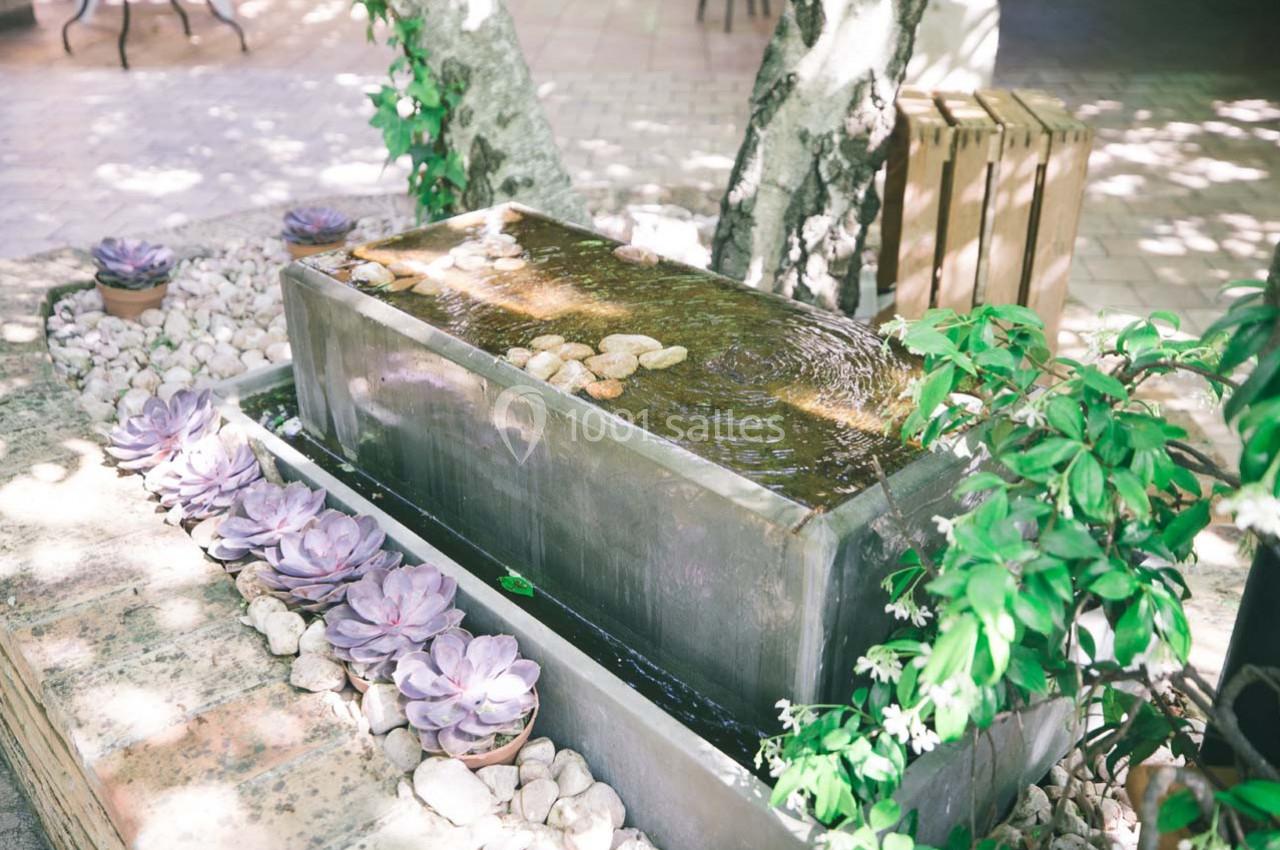 Location salle Gémenos (Bouches-du-Rhône) - Moulin de Gemenos #16 Fontaine rectangulaire en métal entourée de plantes succulentes et de galets, sous des arbres ombragés.