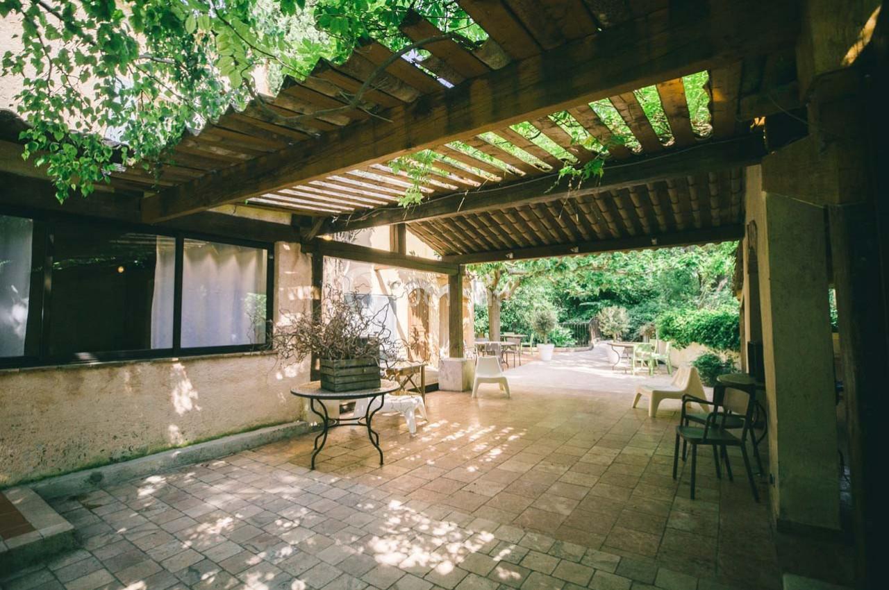 Location salle Gémenos (Bouches-du-Rhône) - Moulin de Gemenos #15 Pergola en bois ombragée sur une terrasse en pierre, entourée de végétation et de mobilier de jardin.