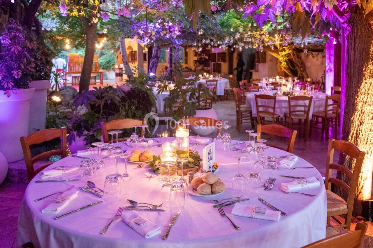 Location salle Gémenos (Bouches-du-Rhône) - Moulin de Gemenos #4 Table ronde dressée pour un dîner en extérieur, entourée de chaises, sous des guirlandes lumineuses et végétation.