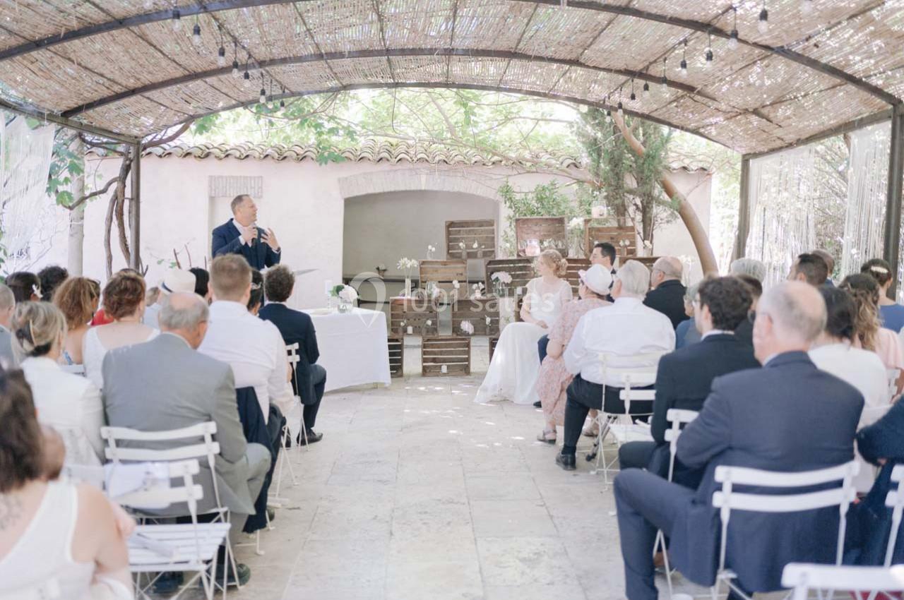 Location salle Gémenos (Bouches-du-Rhône) - Moulin de Gemenos #20 Cérémonie de mariage en extérieur sous une pergola, avec des invités assis et un orateur s'exprimant devant eux.