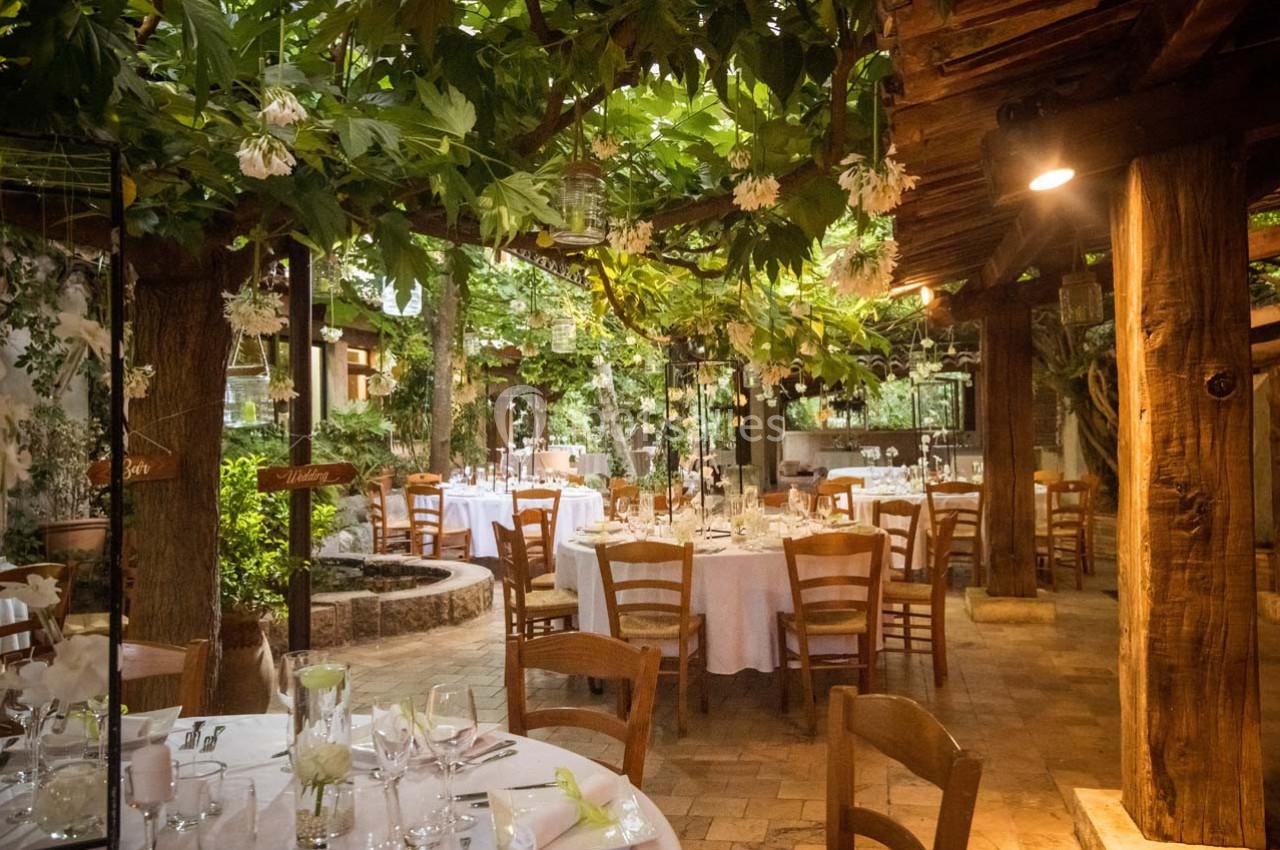 Location salle Gémenos (Bouches-du-Rhône) - Moulin de Gemenos #12 Salle de réception en plein air avec tables dressées, chaises en bois et végétation luxuriante sous un toit partiellement…