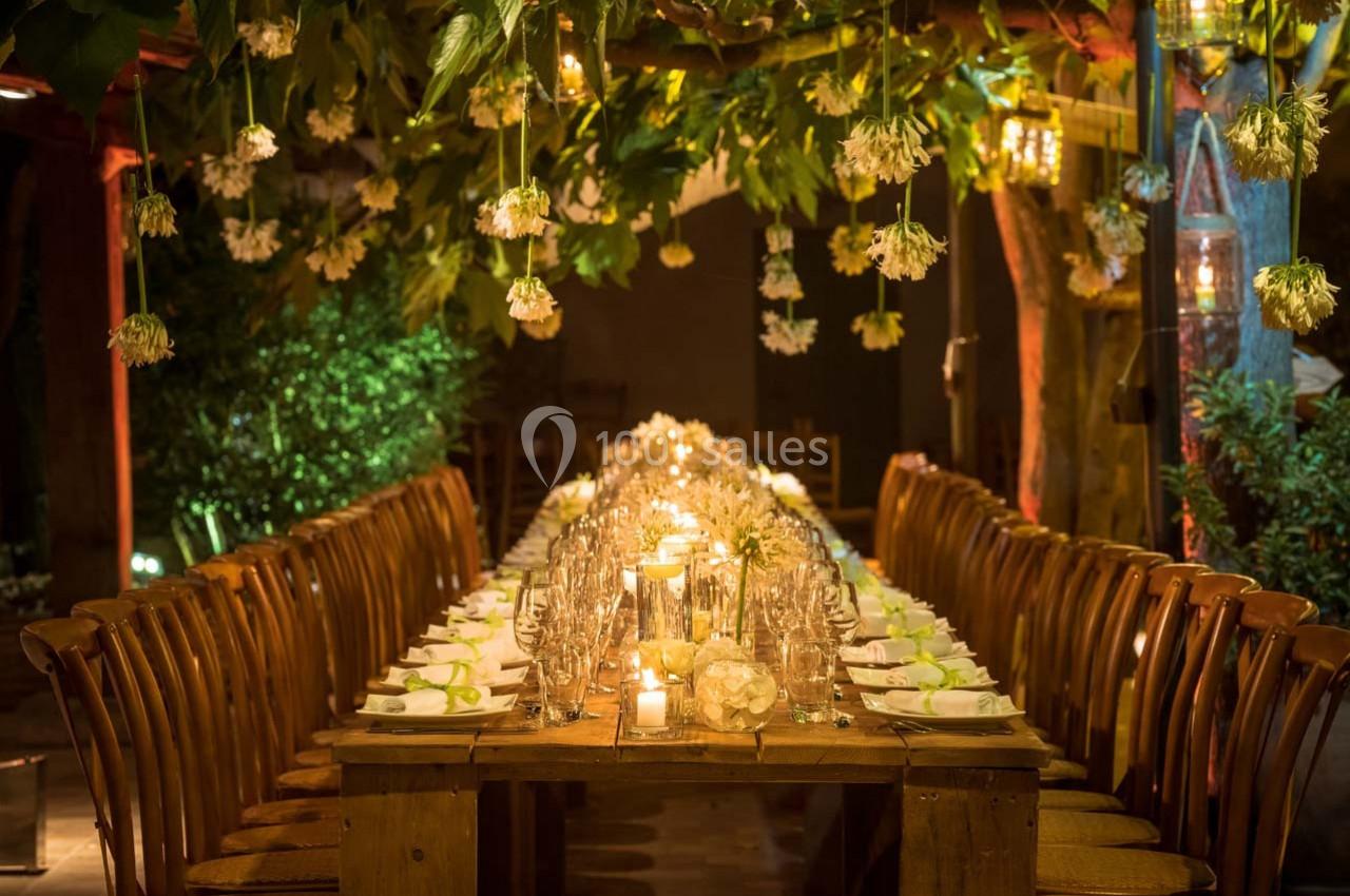 Location salle Gémenos (Bouches-du-Rhône) - Moulin de Gemenos #28 Table en bois décorée pour un dîner, entourée de chaises, avec éclairage chaleureux et fleurs suspendues.