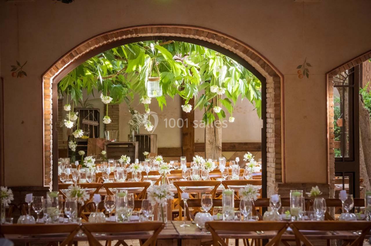 Location salle Gémenos (Bouches-du-Rhône) - Moulin de Gemenos #36 Salle de réception décorée avec des tables en bois, des fleurs blanches et une vue sur un arbre à travers une arche en…