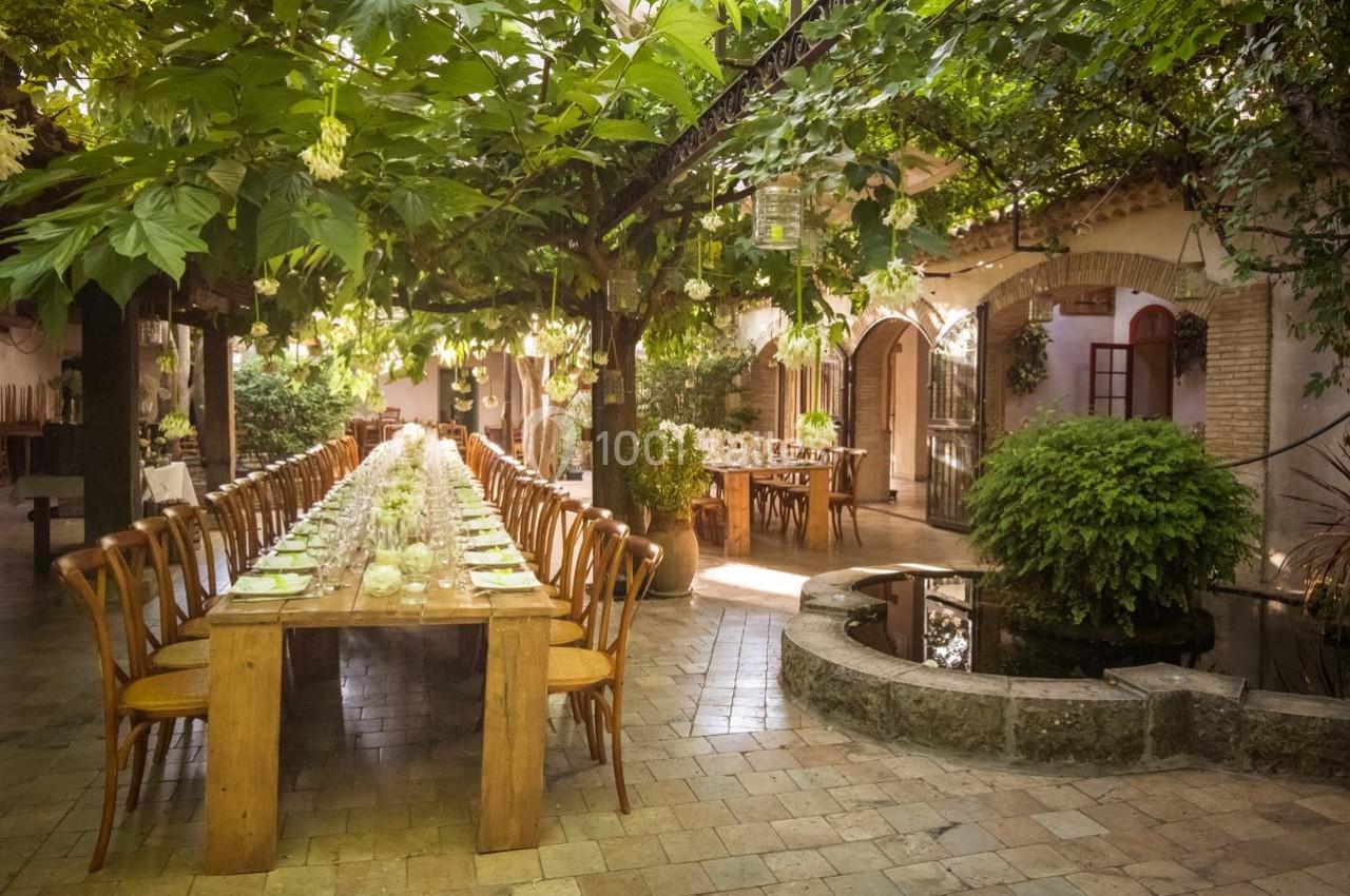 Location salle Gémenos (Bouches-du-Rhône) - Moulin de Gemenos #37 Grande table en bois dressée sous une pergola verdoyante dans une cour pavée avec un petit bassin.