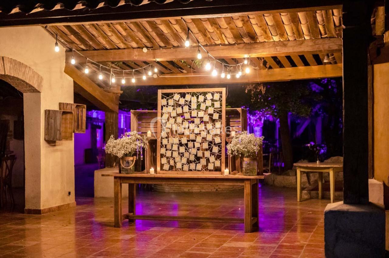 Location salle Gémenos (Bouches-du-Rhône) - Moulin de Gemenos #63 Décoration de mariage en soirée avec guirlandes lumineuses, table en bois et mur orné de photos sous une pergola.