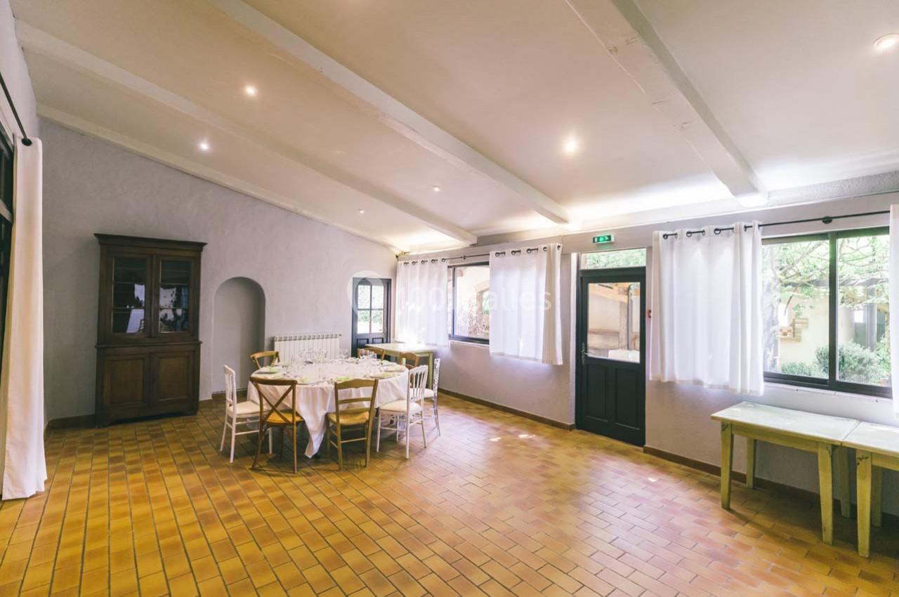 Location salle Gémenos (Bouches-du-Rhône) - Moulin de Gemenos #74 Salle lumineuse avec carrelage en terre cuite, tables dressées, chaises blanches et grandes fenêtres avec rideaux.