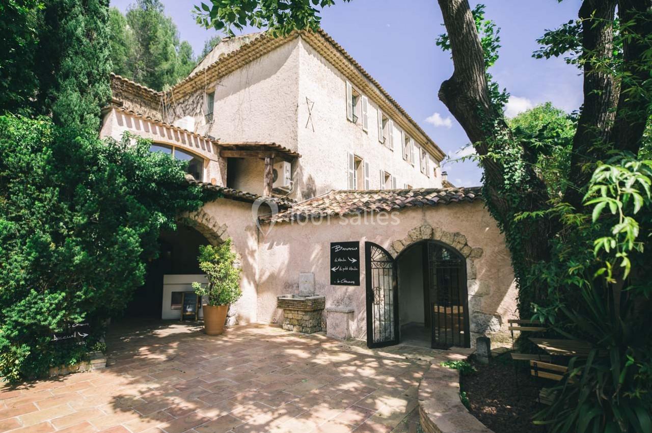 Location salle Gémenos (Bouches-du-Rhône) - Moulin de Gemenos #75 Façade d'un bâtiment en pierre avec toit en tuiles, entouré de végétation et d'une cour pavée ombragée.