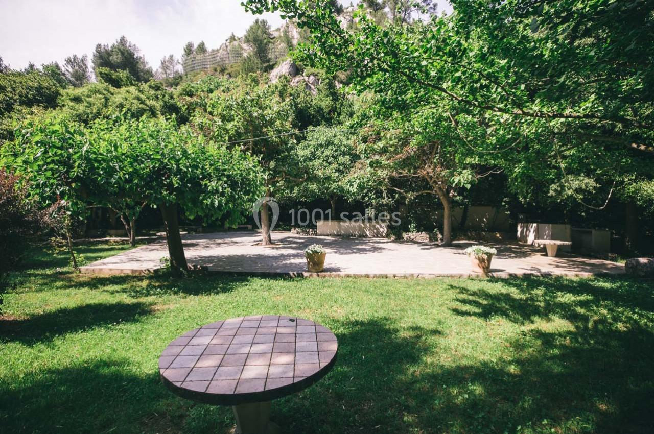 Location salle Gémenos (Bouches-du-Rhône) - Moulin de Gemenos #78 Table ronde en pierre sur une pelouse, entourée d'arbres et d'une terrasse ensoleillée en arrière-plan.
