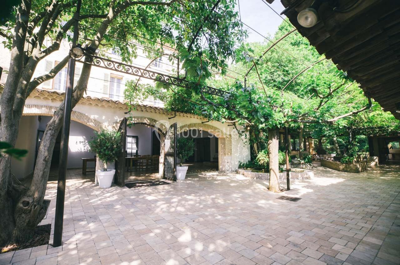 Location salle Gémenos (Bouches-du-Rhône) - Moulin de Gemenos #81 Cour intérieure pavée entourée d'arbres et de végétation, avec une façade en arches et un toit en tuiles.