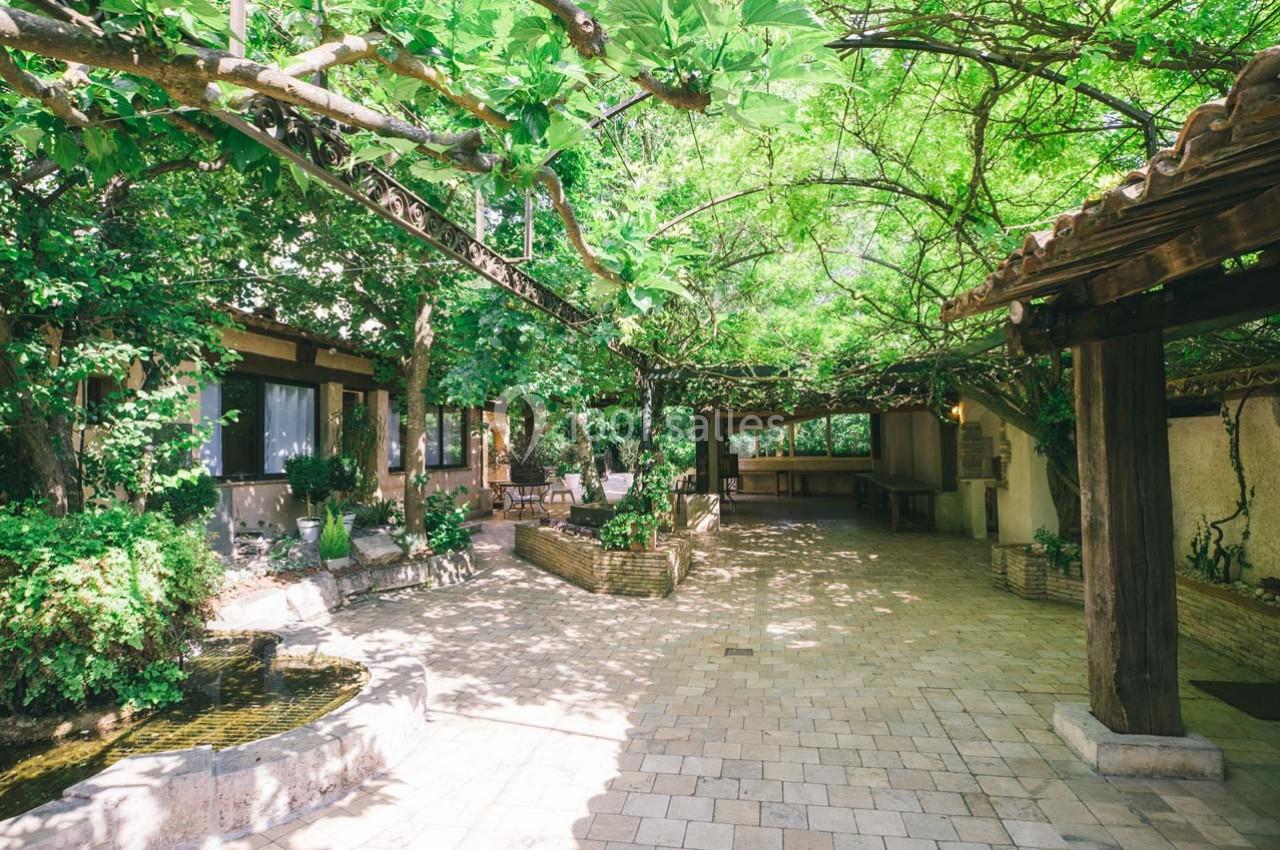 Location salle Gémenos (Bouches-du-Rhône) - Moulin de Gemenos #82 Cour extérieure pavée ombragée par une pergola couverte de végétation, avec un petit bassin et des plantes.