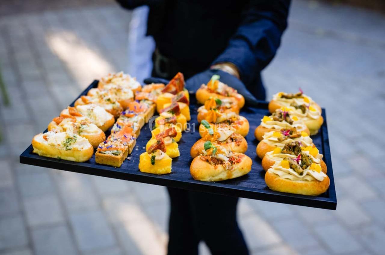 Location salle Gémenos (Bouches-du-Rhône) - Moulin de Gemenos #86 Plateau de canapés variés présentés sur un fond de pavés, tenu par une personne en tenue sombre.