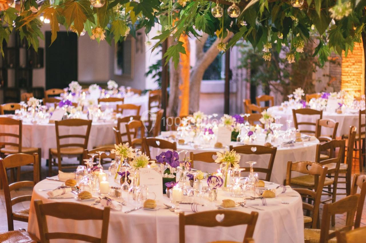 Location salle Gémenos (Bouches-du-Rhône) - Moulin de Gemenos #103 Tables rondes décorées de fleurs, bougies et vaisselle élégante dans un cadre extérieur avec feuillage suspendu.