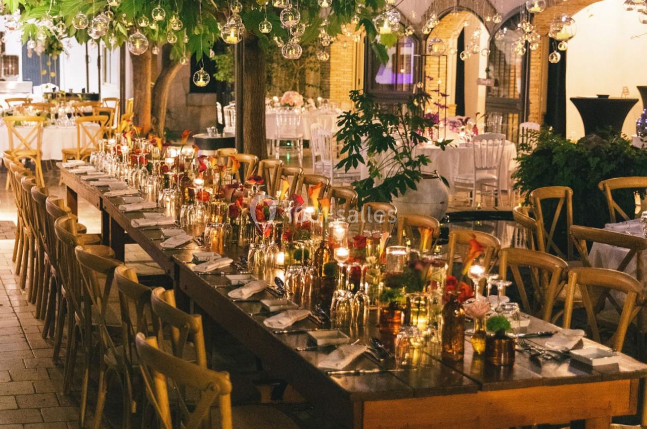 Location salle Gémenos (Bouches-du-Rhône) - Moulin de Gemenos #114 Grande table décorée pour un dîner en extérieur, entourée de chaises en bois, sous des lumières suspendues.
