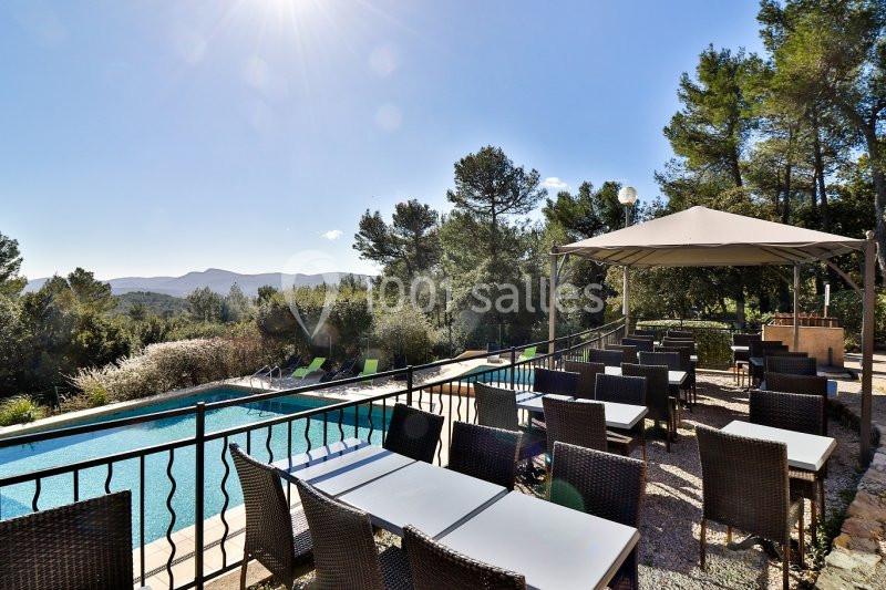 Location salle La Roquebrussanne (Var) - Les Arbousiers #33 Terrasse extérieure avec tables et chaises en rotin, vue sur une piscine, des collines et des arbres sous un ciel ensoleillé.