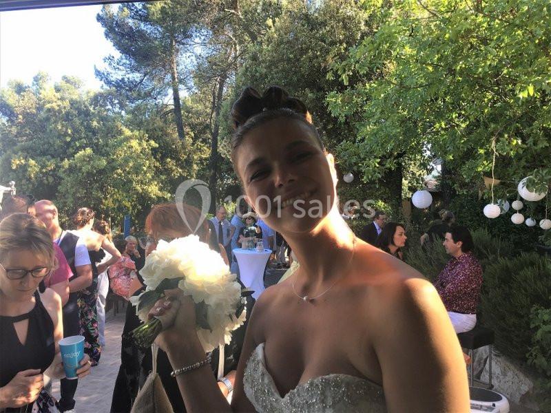 Location salle La Roquebrussanne (Var) - Les Arbousiers #25 Une mariée souriante tenant un bouquet de fleurs blanches lors d'une réception en extérieur, entourée d'invités.