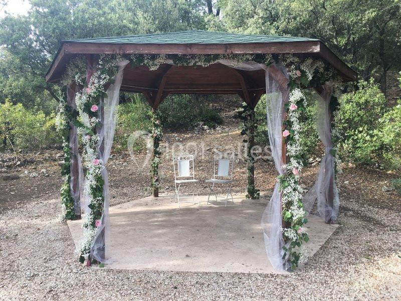 Location salle La Roquebrussanne (Var) - Les Arbousiers #11 Tonnelle en bois décorée de fleurs et voilages, avec deux chaises blanches, située dans un cadre naturel boisé.