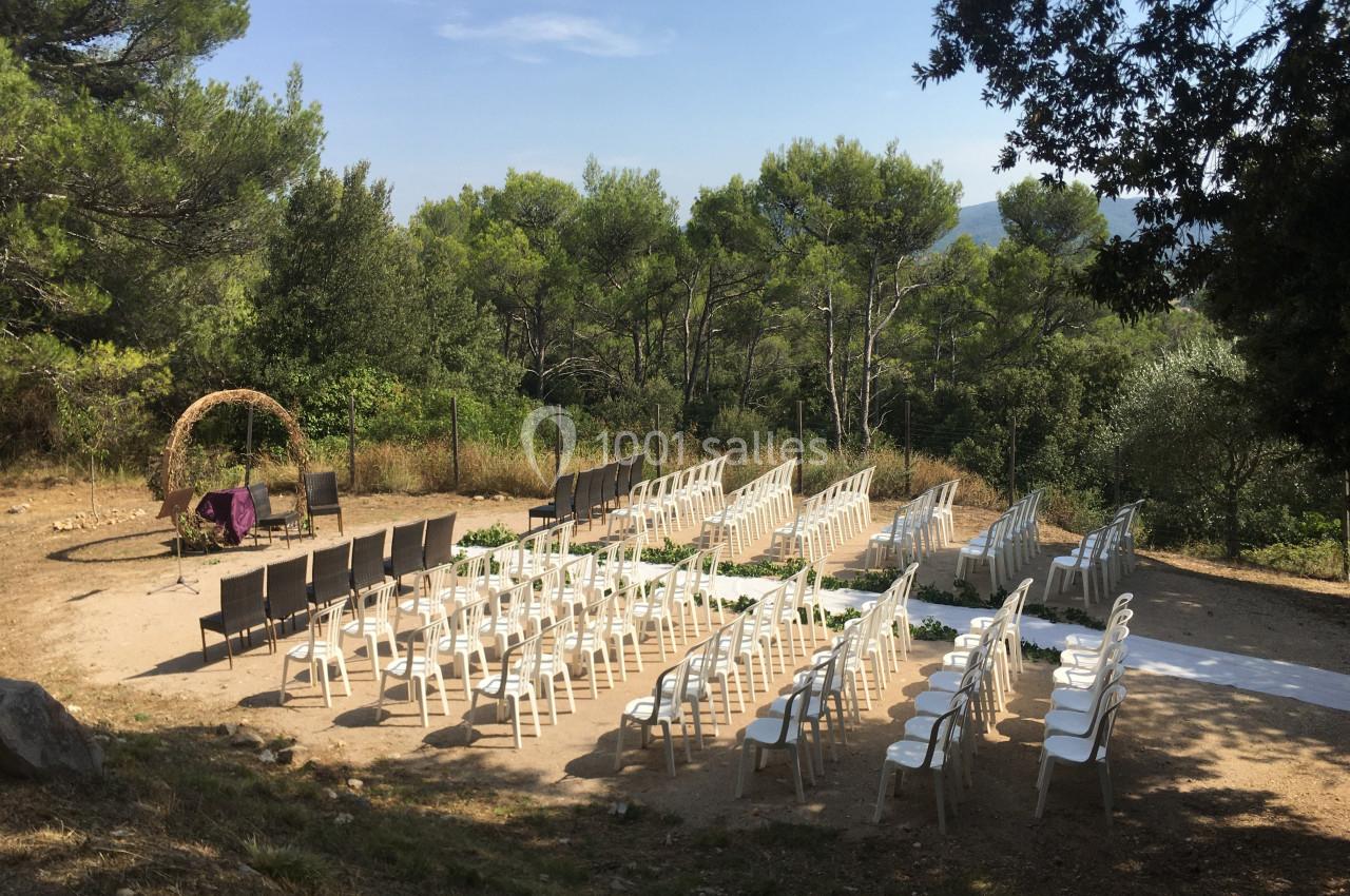 Location salle La Roquebrussanne (Var) - Les Arbousiers #8 Chaises disposées en extérieur sous des arbres, formant une allée centrale, avec une arche décorative en arrière-plan.