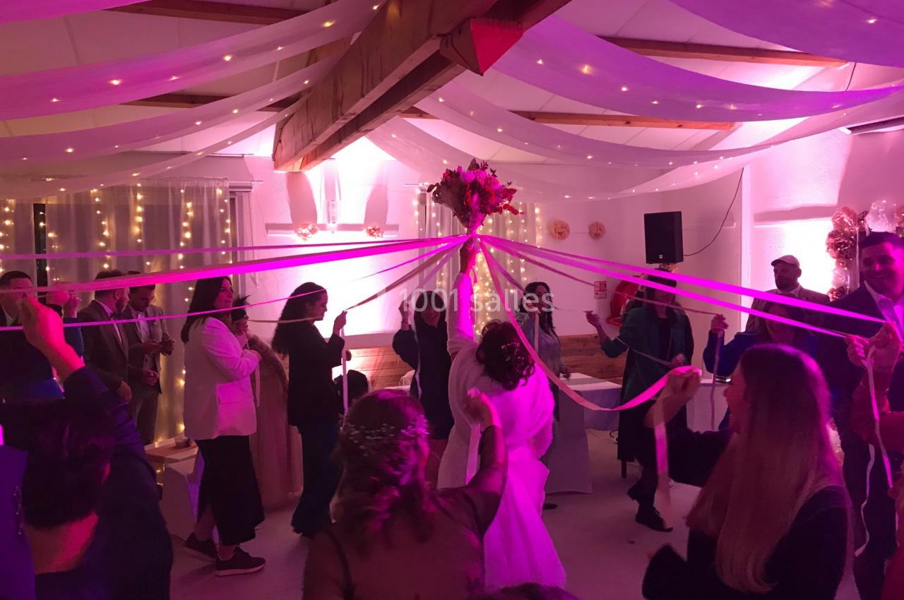 Location salle La Roquebrussanne (Var) - Les Arbousiers #14 Des invités dansent autour de la mariée en tenant des rubans colorés sous un plafond décoré de guirlandes lumineuses.