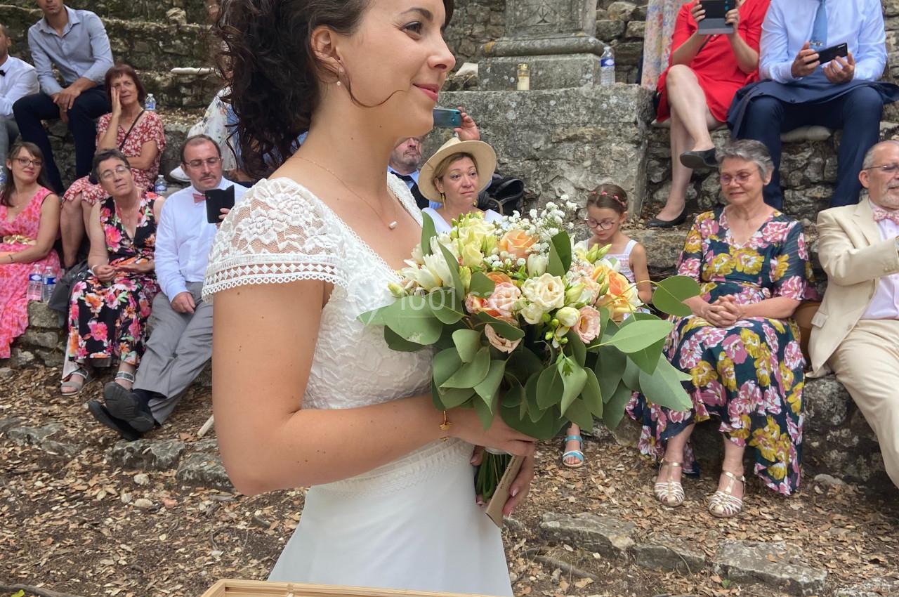 Location salle La Roquebrussanne (Var) - Les Arbousiers #16 Une mariée en robe blanche tient un bouquet de fleurs devant des invités assis en plein air.