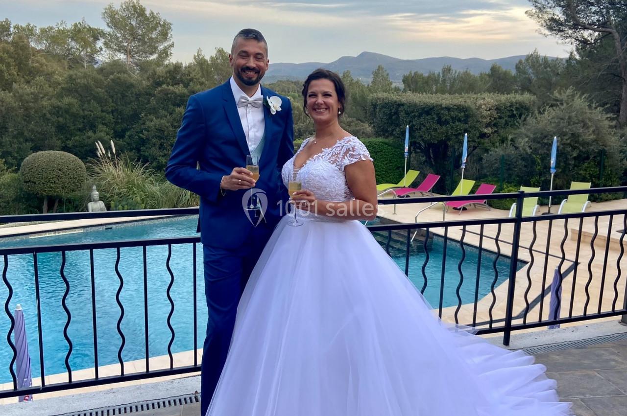 Location salle La Roquebrussanne (Var) - Les Arbousiers #22 Un couple en tenue de mariage pose devant une piscine extérieure avec un paysage arboré en arrière-plan.