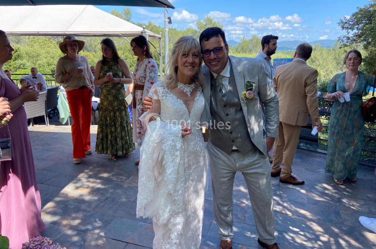 Location salle La Roquebrussanne (Var) - Les Arbousiers #13 Un couple en tenue de mariage sourit sous un ciel bleu, entouré d'invités sur une terrasse extérieure.