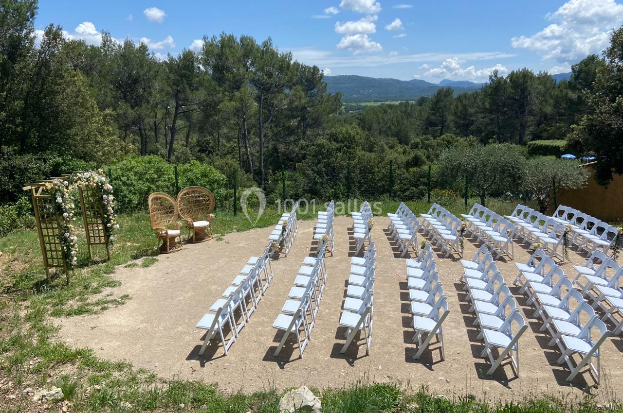 Location salle La Roquebrussanne (Var) - Les Arbousiers #12 Chaises blanches disposées en rangées face à une arche fleurie dans un cadre naturel avec vue sur des collines boisées.