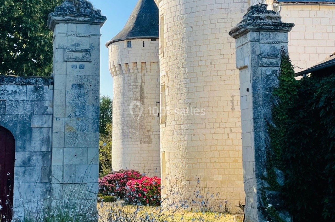 Vue partielle d'un château en pierre blanche avec tours rondes, encadré par des piliers et des fleurs au premier plan.