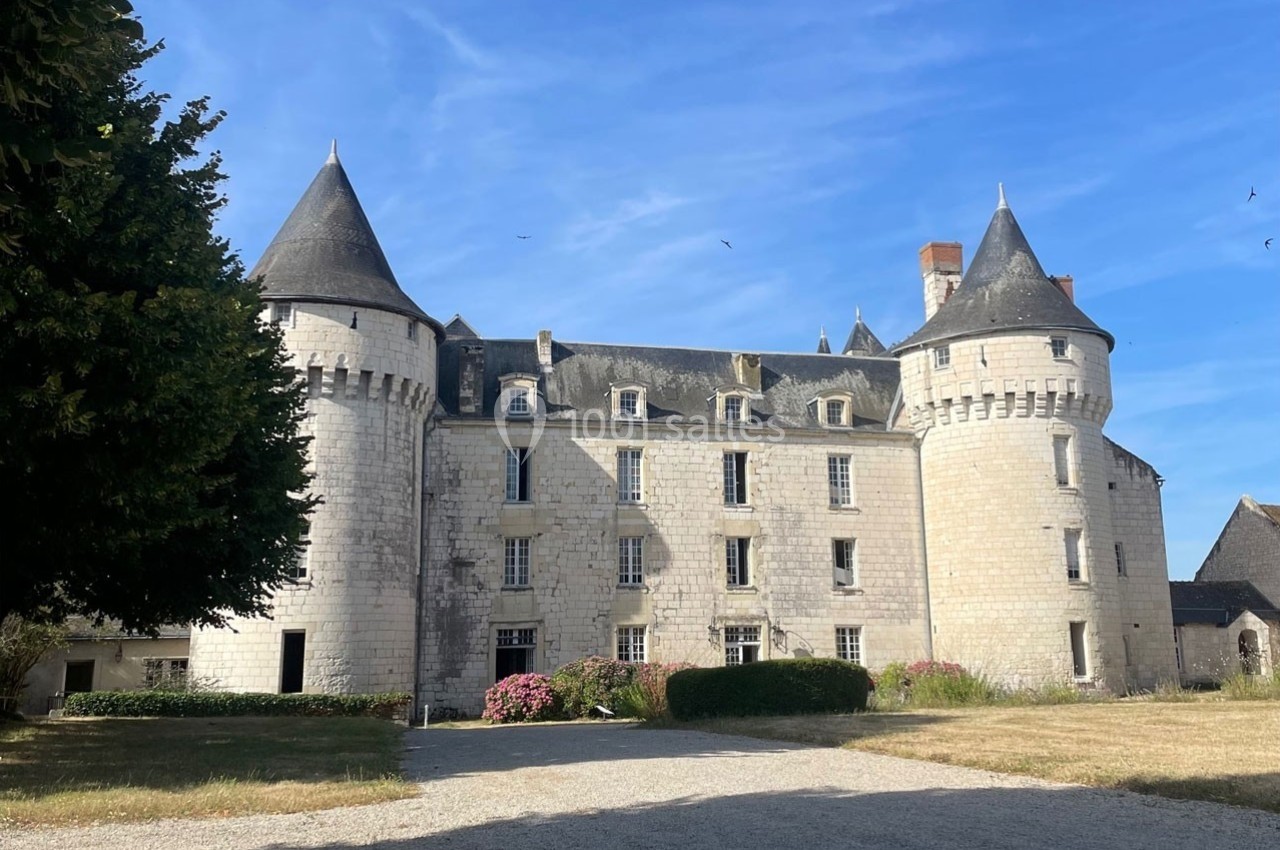 Façade d'un château en pierre blanche avec tours rondes et toit en ardoise, entouré d'une cour et de végétation.