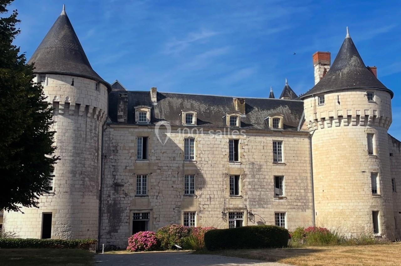 Château en pierre blanche avec tours rondes et toit d'ardoise, entouré de végétation sous un ciel bleu.