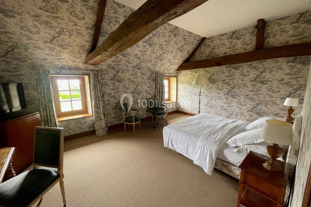 Chambre mansardée avec poutres apparentes, lit double, mobilier classique et fenêtres donnant sur l'extérieur.