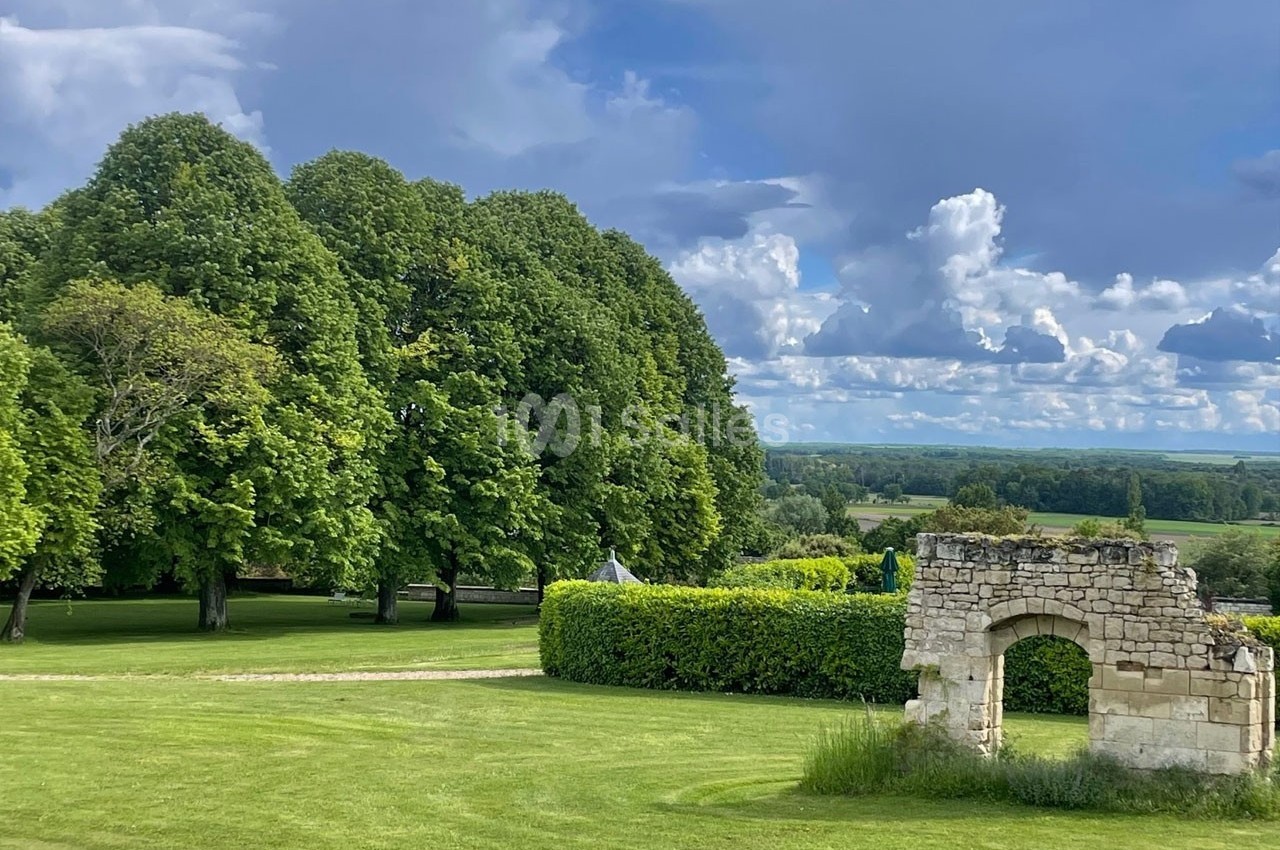 Ruine en pierre dans un parc verdoyant avec pelouse, arbres et ciel nuageux en arrière-plan.