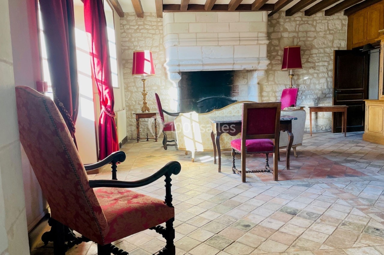 Salle de séjour lumineuse avec cheminée en pierre, meubles anciens et rideaux rouges encadrant des fenêtres.