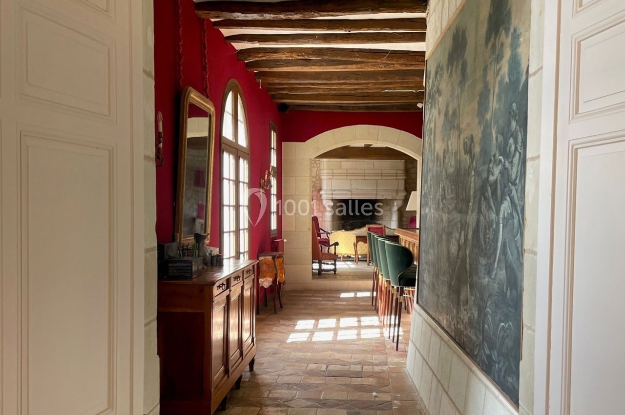 Couloir lumineux avec murs rouges, poutres apparentes, meuble en bois et vue sur un salon avec cheminée en pierre.