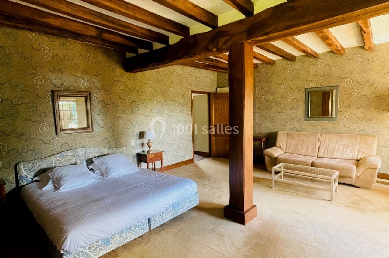 Chambre avec poutres apparentes, lit double, canapé beige, table basse en verre et murs décorés de papier peint.