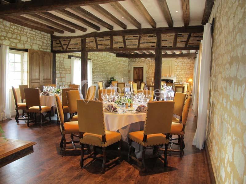 Salle de réception avec murs en pierre, poutres apparentes et tables rondes dressées pour un repas.