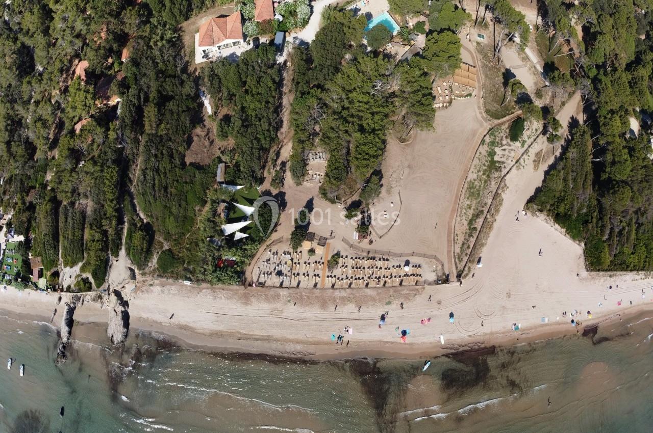 Vue aérienne d'une plage bordée de végétation dense, avec des parasols, des bâtiments et une piscine en arrière-plan.