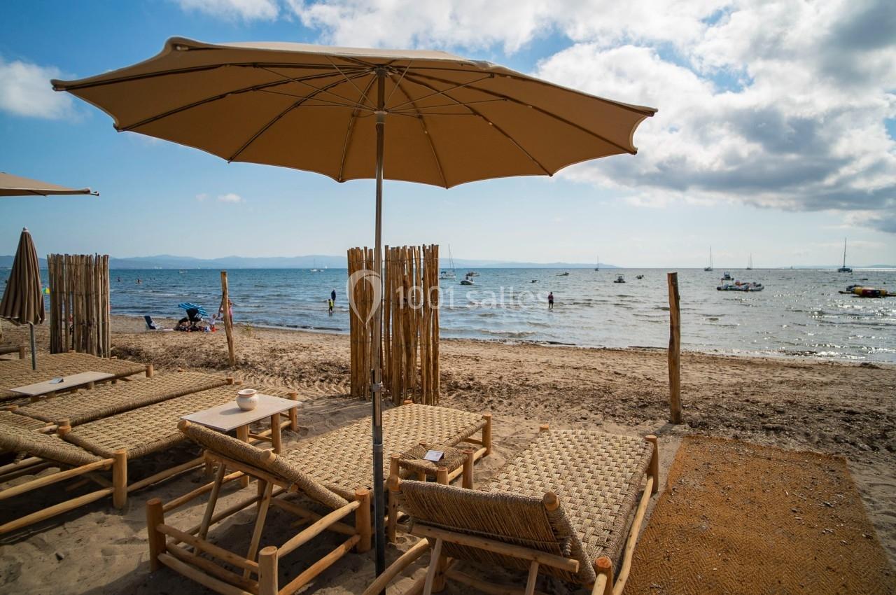 Chaises longues en rotin sous un parasol sur une plage avec vue sur la mer et des bateaux au loin.