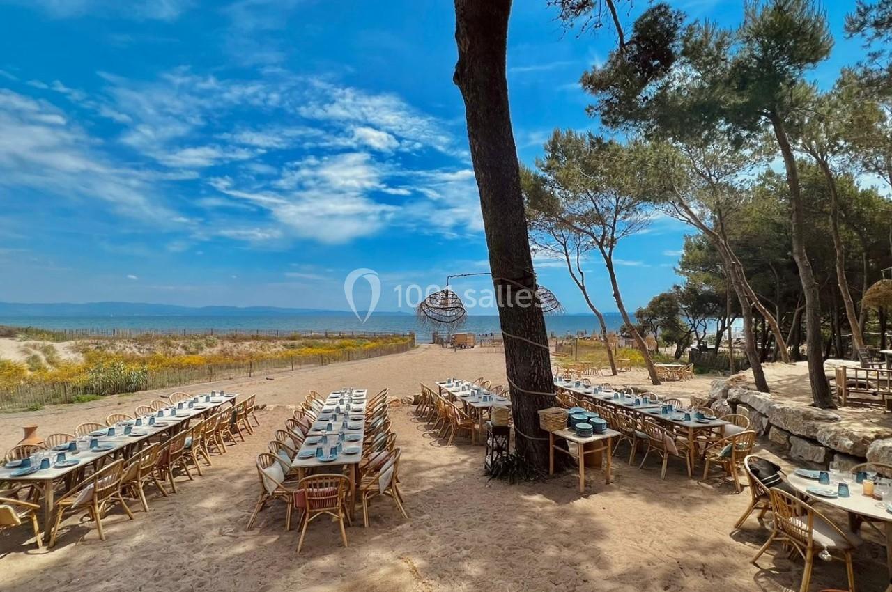 Tables dressées en extérieur sur une plage, entourées de pins, avec vue sur la mer sous un ciel bleu.